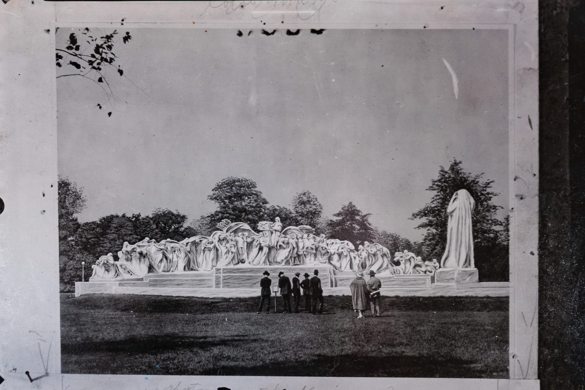 Retouched postcard imaged, five men stand in front of the fountain wearing black, a man and a woman behind, they're all wearing hats.