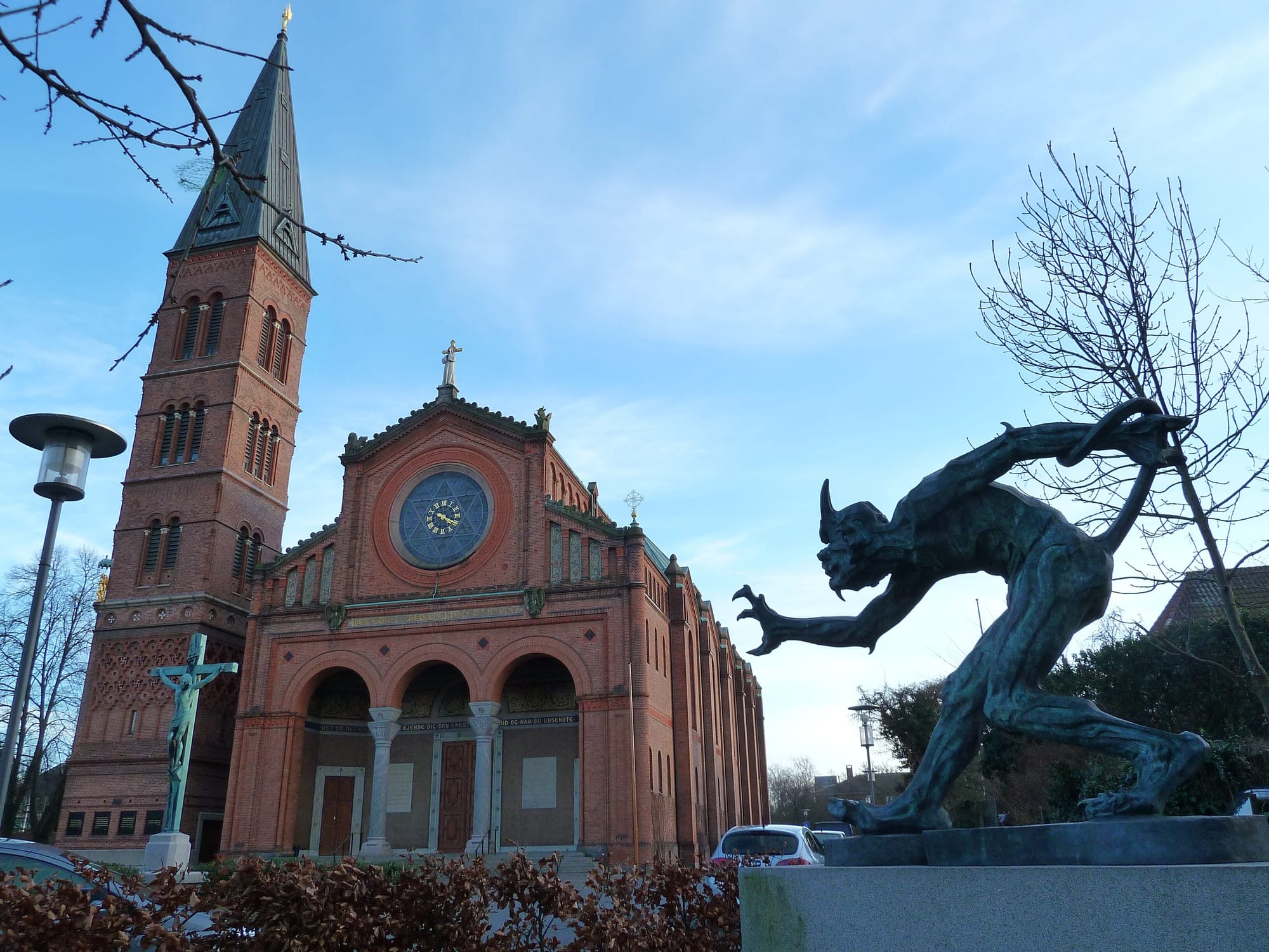 Bronze cast of a troll sculpture standing in front of a pinkish brick church with a big rose window and a campanile