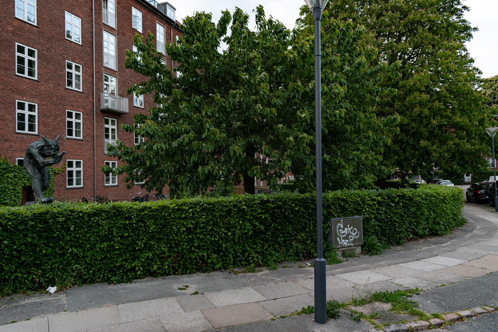 A hedge, a light pole, and a normal brick apartment behind it...but also a creepy statue of a grasping troll on the left.